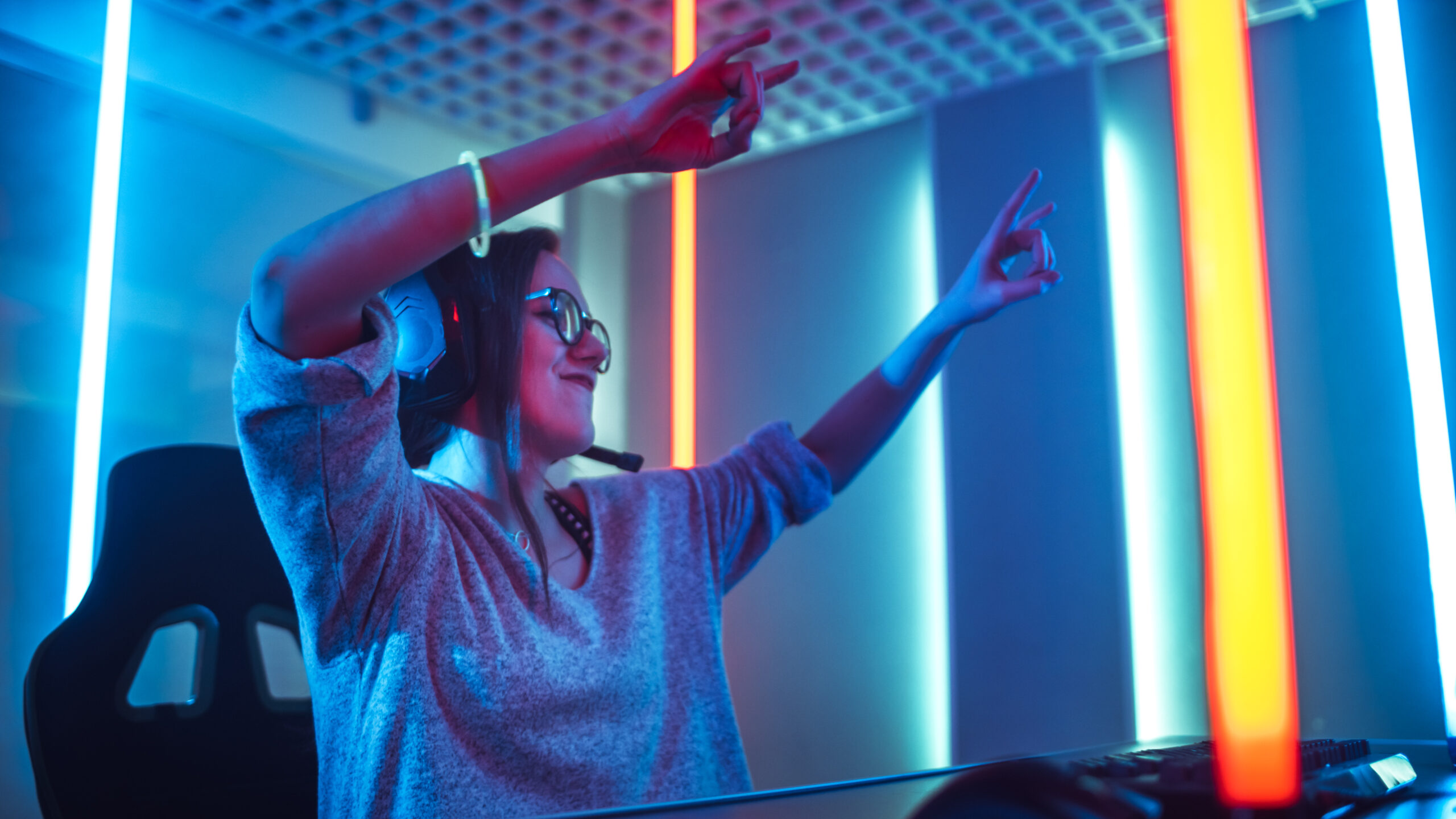 Low Angle Shot of the Beautiful Friendly Pro Gamer Girl Streaming Online Video Game and Winning it, Wearing Headset and Shares with Her Fans. Teenagers Having Fun. Background Cool Neon Retro Colors.