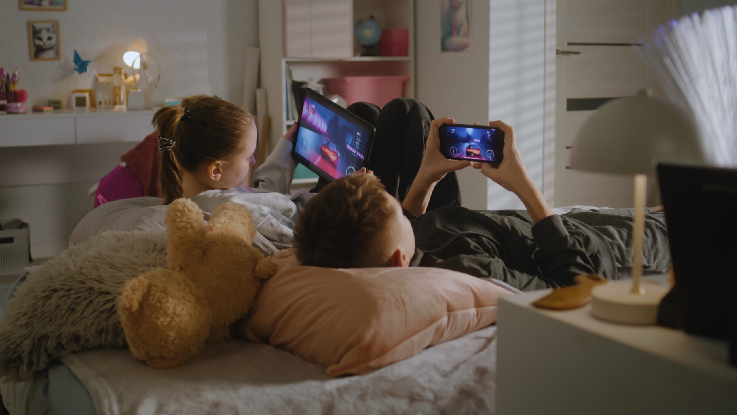 Two teenagers lie on bed in their room and play video games